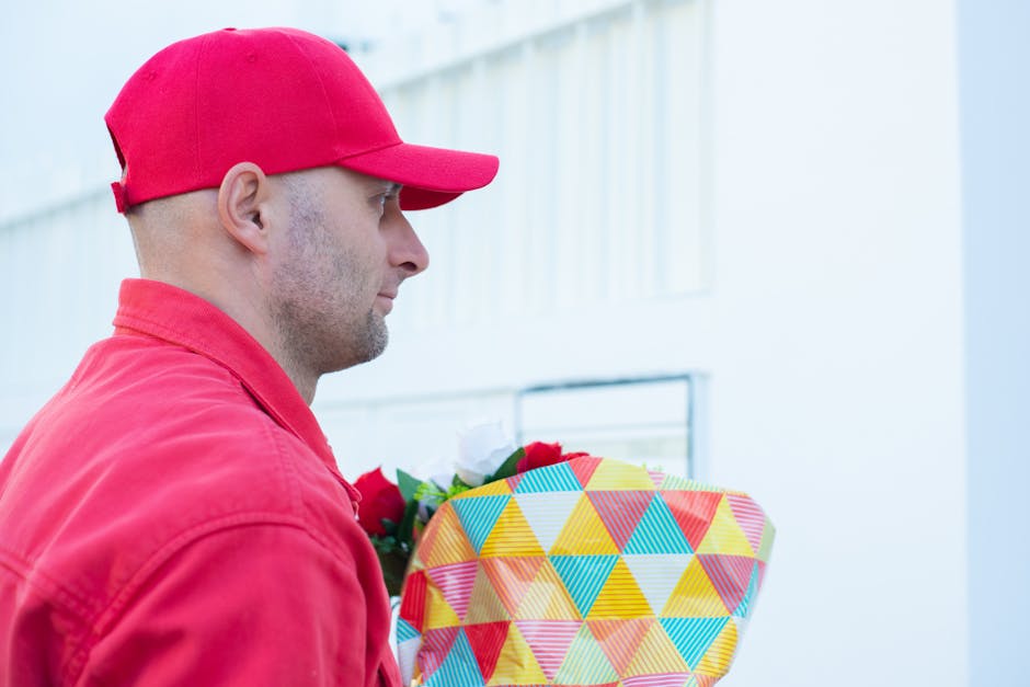 A delivery person dressed in a red cap and red jacket holding a vibrant bouquet of fresh flowers, including white, red, and green foliage, wrapped in colorful geometric patterned paper with shades of 