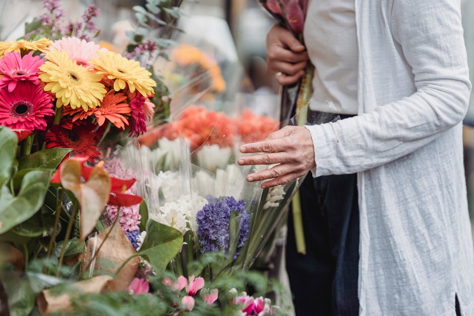 A person wearing a light grey cardigan and dark pants is selecting fresh flowers at a market stall. The arrangement features brightly coloured gerbera daisies in shades of pink, yellow, and orange, al